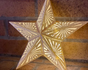 May include: A white, paper star with a geometric design and a string of lights inside. The star is hanging on a brick wall.