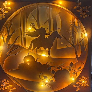 May include: A paper light sculpture featuring a silhouette of a figure riding a horse through a forest with a jack-o-lantern in the background. The sculpture is illuminated with warm white LED lights.