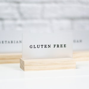 May include: Three white acrylic signs with black text on wooden stands. The signs read "Vegetarian", "Gluten Free", and "Ham".