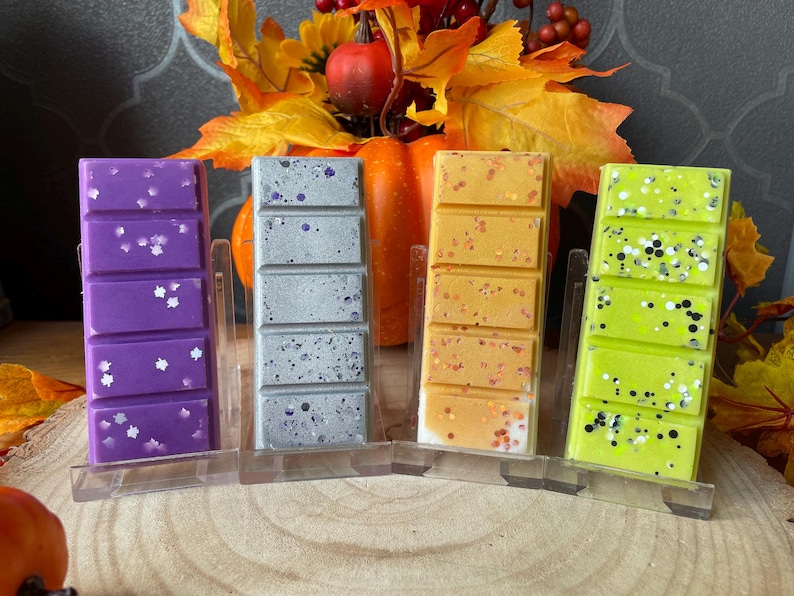 May include: Four rectangular wax melt bars in purple, silver, orange, and lime green, each divided into five segments. The purple bar has white leaf-shaped glitter, the silver has dark speckles, the orange has copper glitter, and the lime green has black and white glitter.