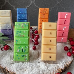 May include: Six rectangular wax melts arranged in clear stands. Colors include white and purple, blue, orange, green with glitter, light yellow with gingerbread man shapes, and pink with white candy cane shapes, all featuring segmented bars.