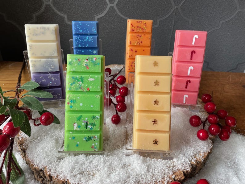 May include: Six rectangular wax melts arranged in clear stands. Colors include white and purple, blue, orange, green with glitter, light yellow with gingerbread man shapes, and pink with white candy cane shapes, all featuring segmented bars.