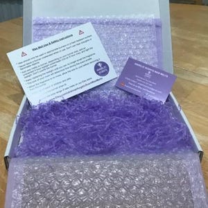 Op de afbeelding: Een open witte doos gevuld met paars cr&ecirc;pepapier en bubbeltjesplastic. Twee kaarten zijn zichtbaar, &eacute;&eacute;n met veiligheidsinstructies en de andere met de tekst "ANGELIC SCENTS WAX MELTS". De doos staat op een houten oppervlak.