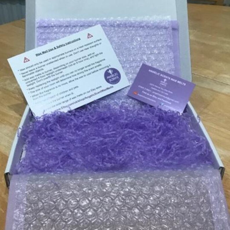 Op de afbeelding: Een open witte doos gevuld met paars cr&ecirc;pepapier en bubbeltjesplastic. Twee kaarten zijn zichtbaar, &eacute;&eacute;n met veiligheidsinstructies en de andere met de tekst "ANGELIC SCENTS WAX MELTS". De doos staat op een houten oppervlak.