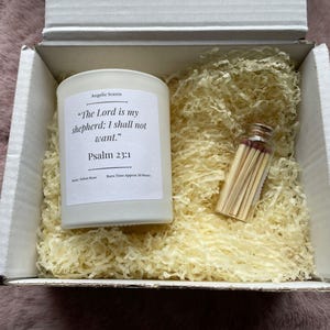 May include: A white candle with the text "The Lord is my shepherd; I shall not want. Psalm 23:1" in a gift box with matches and shredded paper. The candle is labeled "Angelic Scents". The box is white and sits on a pink surface.