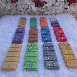 May include: Twelve rectangular wax melts arranged in three rows on a white textured surface. The bars feature various colors including tan, green, orange, purple, blue, red, and gray, decorated with glitter, star shapes, and snowflake accents.