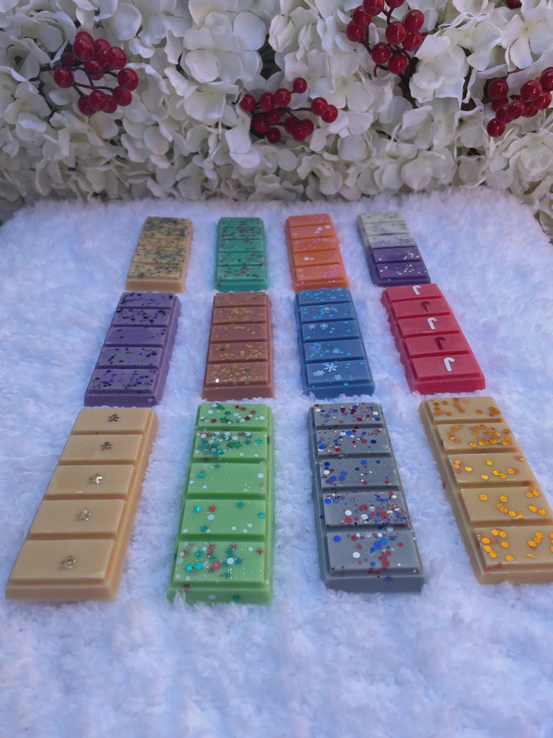 May include: Twelve rectangular wax melts arranged in three rows on a white textured surface. The bars feature various colors including tan, green, orange, purple, blue, red, and gray, decorated with glitter, star shapes, and snowflake accents.