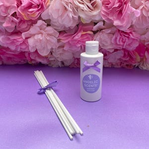 May include: A white bottle of "Angelic Scents" fragrance with a purple bow, next to a bundle of white diffuser sticks tied with a purple ribbon. The background features pink and purple flowers, all set on a purple surface.