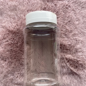 Op de afbeelding: Een transparante plastic pot met een witte deksel. De pot is cilindrisch en leeg, met een brede opening. De deksel is plat en rond. De pot staat op een roze, zachte ondergrond.