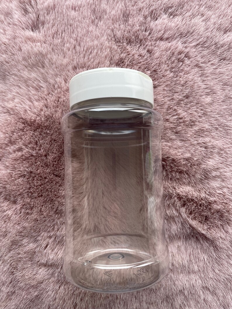 Op de afbeelding: Een transparante plastic pot met een witte deksel. De pot is cilindrisch en leeg, met een brede opening. De deksel is plat en rond. De pot staat op een roze, zachte ondergrond.
