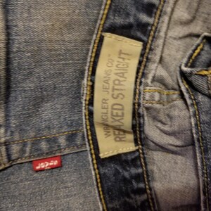May include: Close-up of blue denim jeans with a red tag and a beige tag. The red tag has white text. The beige tag reads "Wrangler Jeans Co." and "Relaxed Straight".
