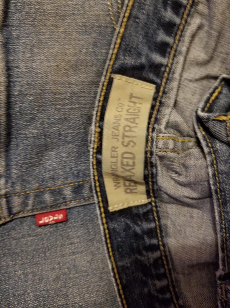 May include: Close-up of blue denim jeans with a red tag and a beige tag. The red tag has white text. The beige tag reads "Wrangler Jeans Co." and "Relaxed Straight".