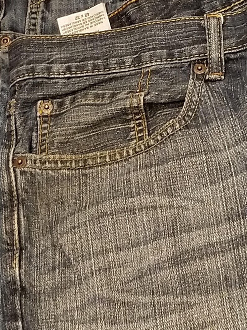 May include: Close-up of blue denim jeans with a classic five-pocket design. The jeans feature a button and zipper closure, belt loops, and rivets. The tag reads "42 x 32". The denim has a worn, faded appearance.