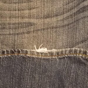 May include: Close-up of denim fabric showing a seam with a frayed edge. The denim has a faded, worn appearance with a mix of light and dark blue tones. The stitching is visible along the seam.