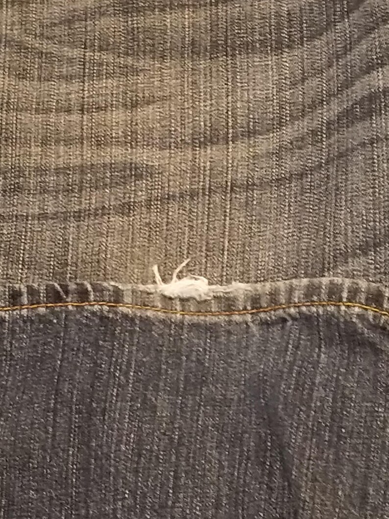 May include: Close-up of denim fabric showing a seam with a frayed edge. The denim has a faded, worn appearance with a mix of light and dark blue tones. The stitching is visible along the seam.