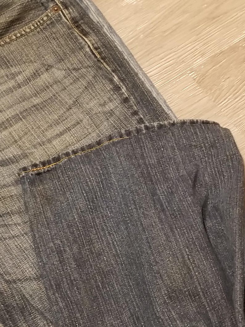 May include: Close-up of denim jeans in shades of gray and blue. The jeans are folded, showing the texture of the fabric and the stitching details. The image highlights the worn look of the denim.
