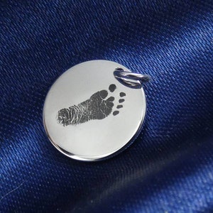 Könnte beinhalten: Ein runder Anhänger aus Silber mit einem eingravierten Baby-Fußabdruck.