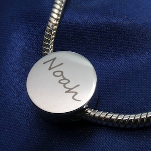 Könnte beinhalten: Ein silbernes Armband mit einem runden silbernen Anhänger, auf dem "Noah" eingraviert ist.