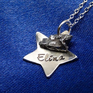 Könnte beinhalten: Eine silberne Halskette mit einem sternförmigen Anhänger. Der Anhänger trägt den Namen "Elina" und ein kleines Stück Meteoriten auf der Oberseite.
