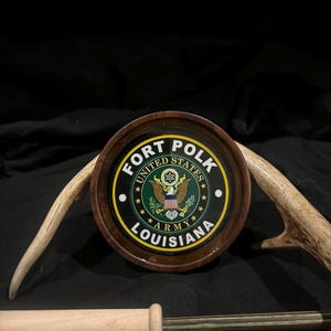 May include: A round wooden plaque with a green and gold emblem featuring the United States Army seal and the text "FORT POLK LOUISIANA".