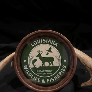 May include: A round wooden coaster featuring the Louisiana Department of Wildlife & Fisheries logo. The logo includes a deer, fish, and flying ducks in a green and white design, with the text "Louisiana" and "Wildlife & Fisheries".