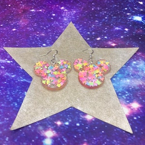 May include: A pair of dangle earrings shaped like Mickey Mouse heads. The earrings are made of clear resin with colorful glitter embedded inside. The glitter is in various shades of pink, blue, yellow, and green.