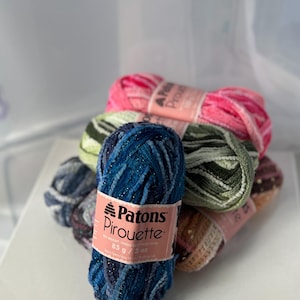 May include: A collection of Patons Pirouette yarn skeins in various colors, including blue, pink, green, and brown. Each skein is labeled with the brand name and weight, 85 g / 3 oz. The yarn has a textured appearance, suitable for knitting and crochet projects.