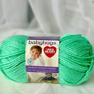 May include: A skein of mint green Baby Hugs yarn by Red Heart. The label features a baby's photo and the words "Baby Hugs" and "Red Heart". The yarn is medium weight, 4.5 oz / 127 g.