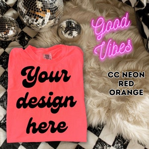 Op de afbeelding: Een felroze T-shirt met de tekst "Your design here" in een retro-lettertype. Het shirt ligt op een geruite achtergrond met discoballen en een pluche wit tapijt. De tekst "Good Vibes" staat in roze neonletters boven het tapijt. De tekst "CC Neon Red Orange" staat onder het tapijt.