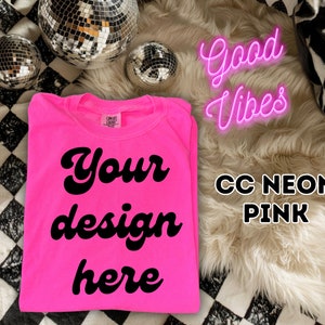 Op de afbeelding: Een felroze T-shirt met de tekst "Your design here" in zwart. Het shirt ligt op een zwart-wit geblokt oppervlak met een pluche wit tapijt en een neonroze bord met de tekst "Good Vibes".