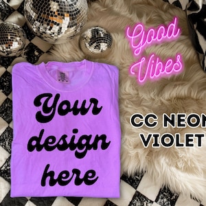 Op de afbeelding: Een paars T-shirt met de tekst "Your design here" in zwart. Het shirt ligt op een zwart-wit geblokte achtergrond met een pluche wit tapijt en discoballen. De tekst "Good Vibes" is in roze neonletters en de tekst "CC Neon Violet" is in zwarte letters.