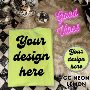 Op de afbeelding: Een neon-geel T-shirt met de tekst "Your design here" in zwart. Het shirt ligt op een wit, pluche oppervlak met een zilveren cirkel waarop "Your design here" en de tekst "CC NEON LEMON" staat.