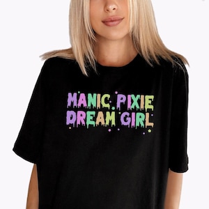 Puede incluir: Camiseta negra con el texto "MANIC PIXIE DREAM GIRL" en colores pastel que gotean.
