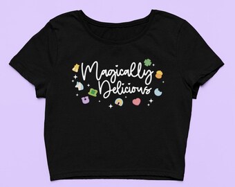 May include: Black cropped t-shirt with the text "Magically Delicious" in white script. The shirt features a variety of colorful cartoon images, including a unicorn, a pot of gold, a horseshoe, a rainbow, and a heart.