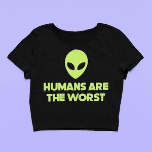 Könnte beinhalten: Schwarzes Crop-Top mit einem neongrünen Alien-Kopf und dem Text "HUMANS ARE THE WORST". Das Shirt hat einen Rundhalsausschnitt und kurze Ärmel. Die Grafik ist mittig auf der Vorderseite platziert.