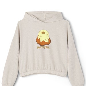 Op de afbeelding: Een licht beige hoodie met een afbeelding van een bruine en gele kaneelrol met de tekst "SWEETROLL" eronder.