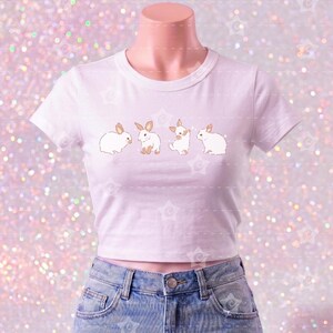 May include: A white short-sleeved crop top featuring a horizontal row of four small brown and white rabbit illustrations across the chest, paired with light blue denim shorts that have a distressed, frayed hemline and visible pocket lining.