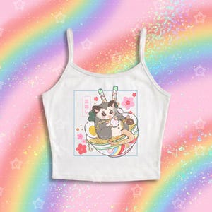 May include: A white spaghetti strap crop top featuring a graphic illustration of a gray and white opossum eating ramen from a bowl with chopsticks. The design includes pink cherry blossoms, a hard-boiled egg, and Japanese text in a blue square frame.