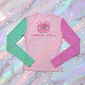 May include: A pink mesh long-sleeved top featuring a cartoon character and the text "Leave me a loan." The sleeves are green and purple. The top has a frilled neckline.