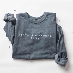 May include: A dark gray sweatshirt with the text "Sorry. I'm awkward. Sorry." in white. The long sleeves are rolled up. The sweatshirt is displayed on a white surface with star-shaped decorations.