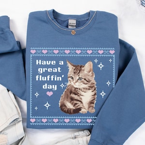 Op de afbeelding: Een blauw sweatshirt met een gepixelde afbeelding van een kat en de tekst "Have a great flufffin' day". Het sweatshirt heeft een patroon van roze harten en sterren.