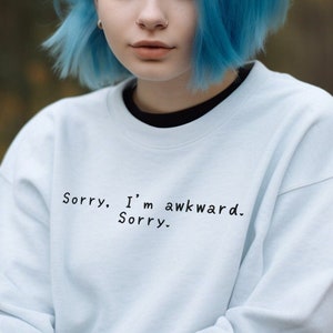 May include: A light blue sweatshirt with the text "Sorry. I'm awkward. Sorry." in black. The sweatshirt has a crew neck and long sleeves. The person in the image has blue hair.