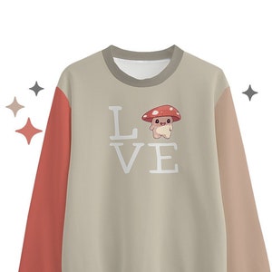 以下が含まれることがあります： 前面に赤と白のキノコのグラフィックと「LOVE」の文字がプリントされたベージュのトレーナー。トレーナーは左側に赤い長袖、右側にベージュの長袖が付いています。