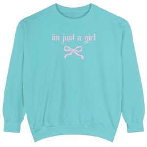 Op de afbeelding: Een turquoise blauwe trui met de tekst "i'm just a girl" in roze letters en een roze strik.