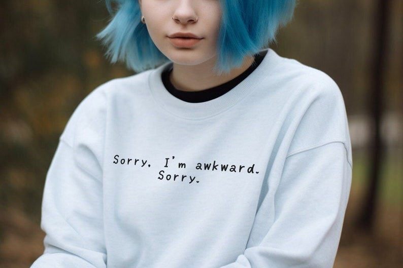 May include: A light blue sweatshirt with the text "Sorry. I'm awkward. Sorry." in black. The sweatshirt has a crew neck and long sleeves. The person in the image has blue hair.