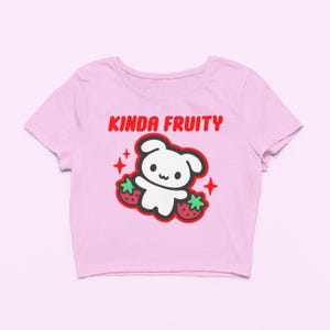 Op de afbeelding: Een lichtroze crop top met een ronde halslijn. De shirt heeft een afbeelding van een witte cartoonbeer met twee aardbeien en de tekst "KINDA FRUITY" in het rood. Het ontwerp bevat rode steraccenten.