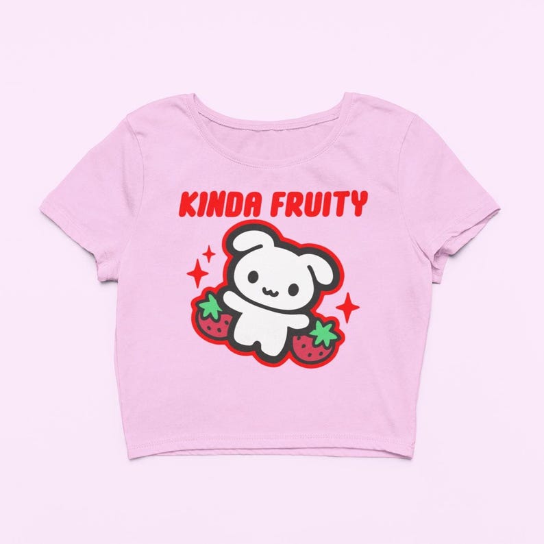 Op de afbeelding: Een lichtroze crop top met een ronde halslijn. De shirt heeft een afbeelding van een witte cartoonbeer met twee aardbeien en de tekst "KINDA FRUITY" in het rood. Het ontwerp bevat rode steraccenten.