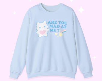 Puede incluir: Sudadera azul claro con la frase "ARE YOU MAD AT ME?" y un oso de dibujos animados con un corazón rosa. Un arcoíris y estrellas también están presentes en el diseño. La sudadera tiene mangas largas.