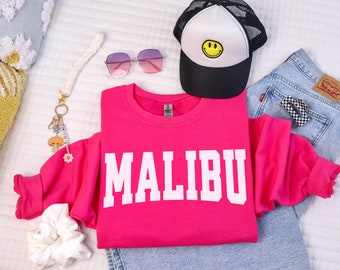 Op de afbeelding: Een roze sweatshirt met het woord "MALIBU" in wit op de voorkant. Het sweatshirt ligt op een wit oppervlak met een zonnebril, een witte scrunchie, een sleutelhanger en een trucker cap met een smiley.