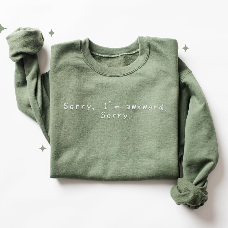 May include: Sage green sweatshirt with the embroidered text "Sorry. I'm awkward. Sorry." The casual crew neck top has rolled-up sleeves and is made of a soft material. The sweatshirt is a comfortable and stylish choice for everyday wear.